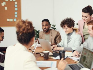 A diverse group of professionals collaborating around a table with laptops, papers, and coffee cups in a modern office, with a corkboard in the background, highlighting real visa sponsorship job success stories.