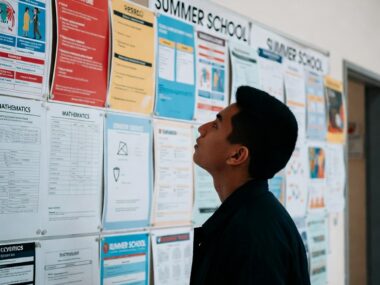 A thoughtful young student stands in front of a wall covered with colorful course posters and summer school flyers, carefully reading each one while deciding his future path.