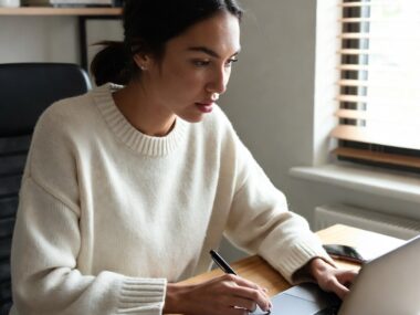 Young female international student sitting at her desk with laptop, notebook, and calculator, carefully tracking expenses and managing finances while studying abroad