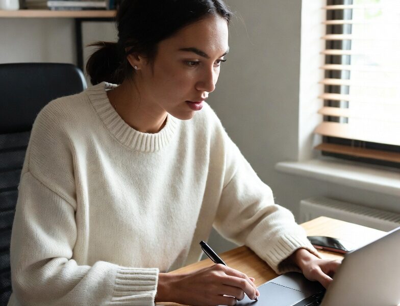 Young female international student sitting at her desk with laptop, notebook, and calculator, carefully tracking expenses and managing finances while studying abroad