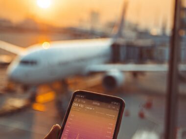 A traveler checking flight prices on a smartphone while waiting at an airport terminal, searching for cheap flights in 2026.